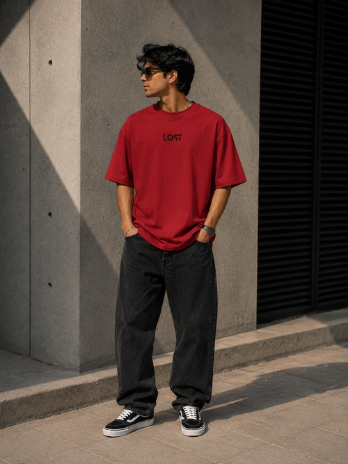 Person wearing a red t-shirt with LOST text, black pants, and black sneakers standing against a gray wall.